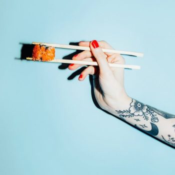 tattooed hand holding a piece of sushi with chopsticks against a blue background